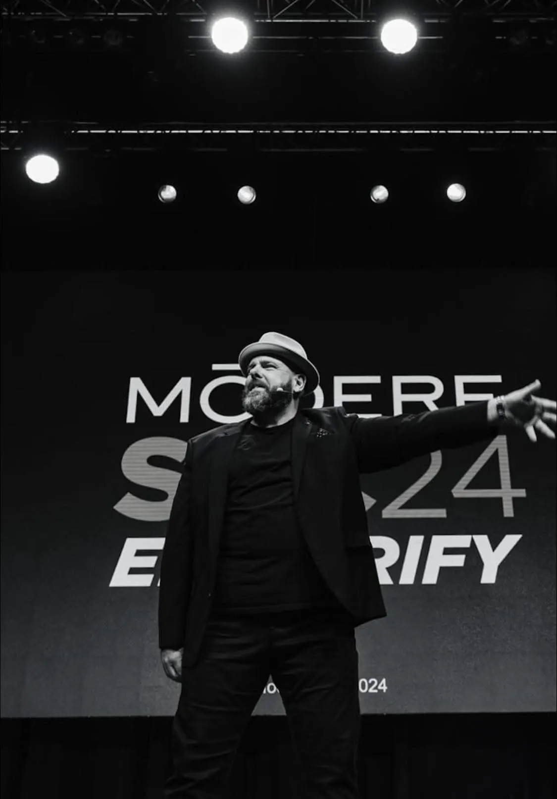 Black and white photo of Brett Davis on stage in a fedora and black jacket, arm stretched out toward the audience