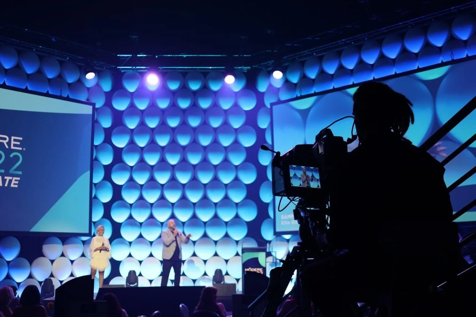 Brett co-hosting on a lit conference stage, filmed by a camera operator in the foreground