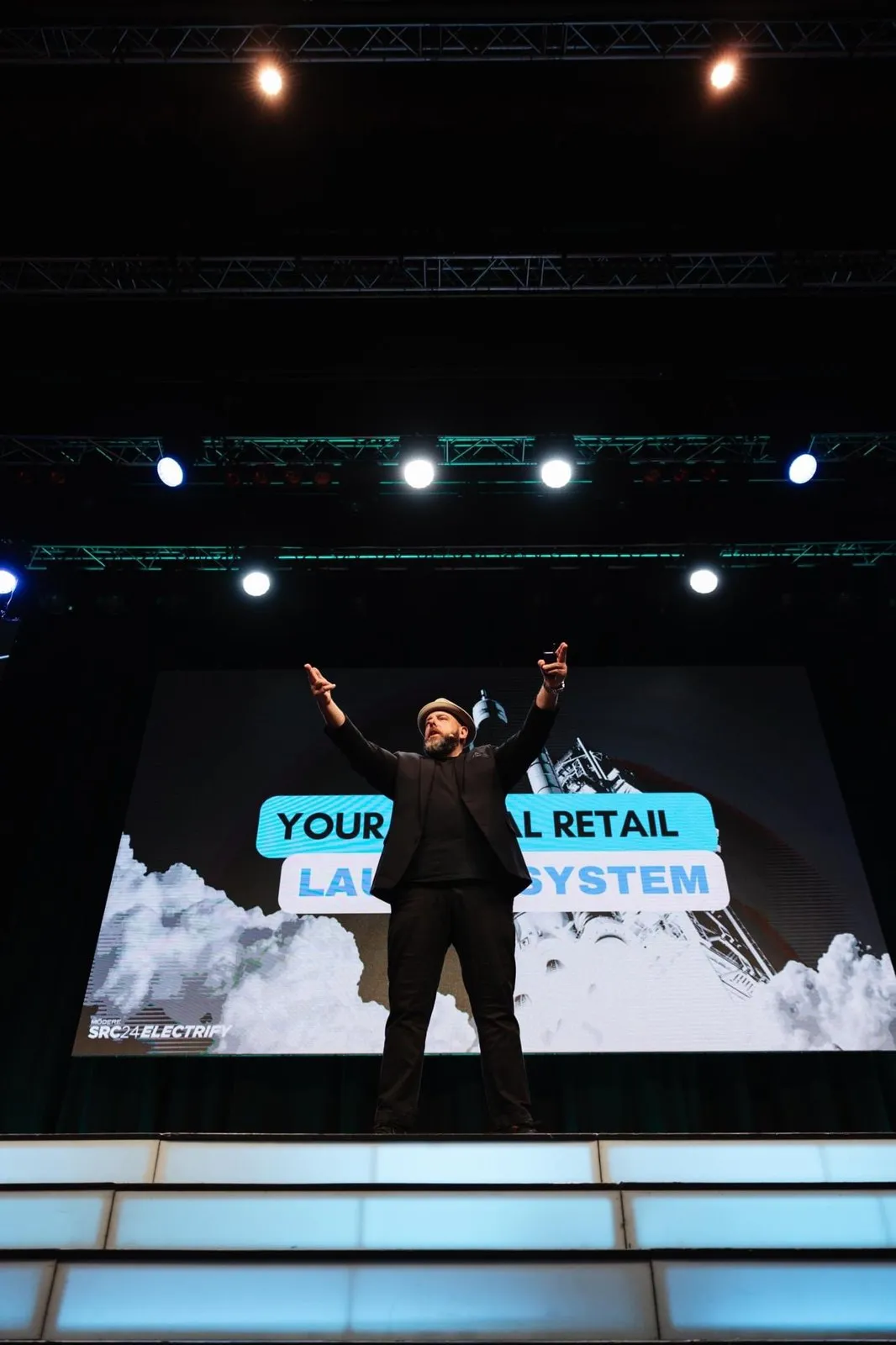 Brett on a large stage with both arms raised under the lighting rig